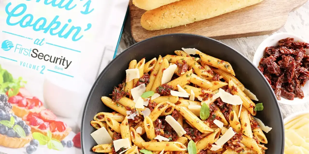 Makaron penne z suszonymi pomidorami, parmezanem i bazylią w czarnej misce, obok pieczywa i książki kucharskiej.