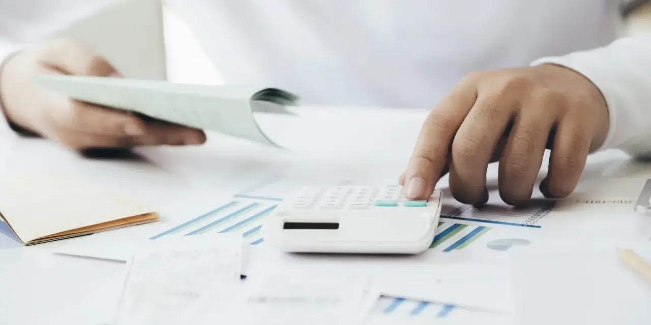 Manos calculando con calculadora blanca sobre gráficos y papeles. Una mano sostiene un talonario, la otra opera la calculadora. Proceso de factura simplificada.