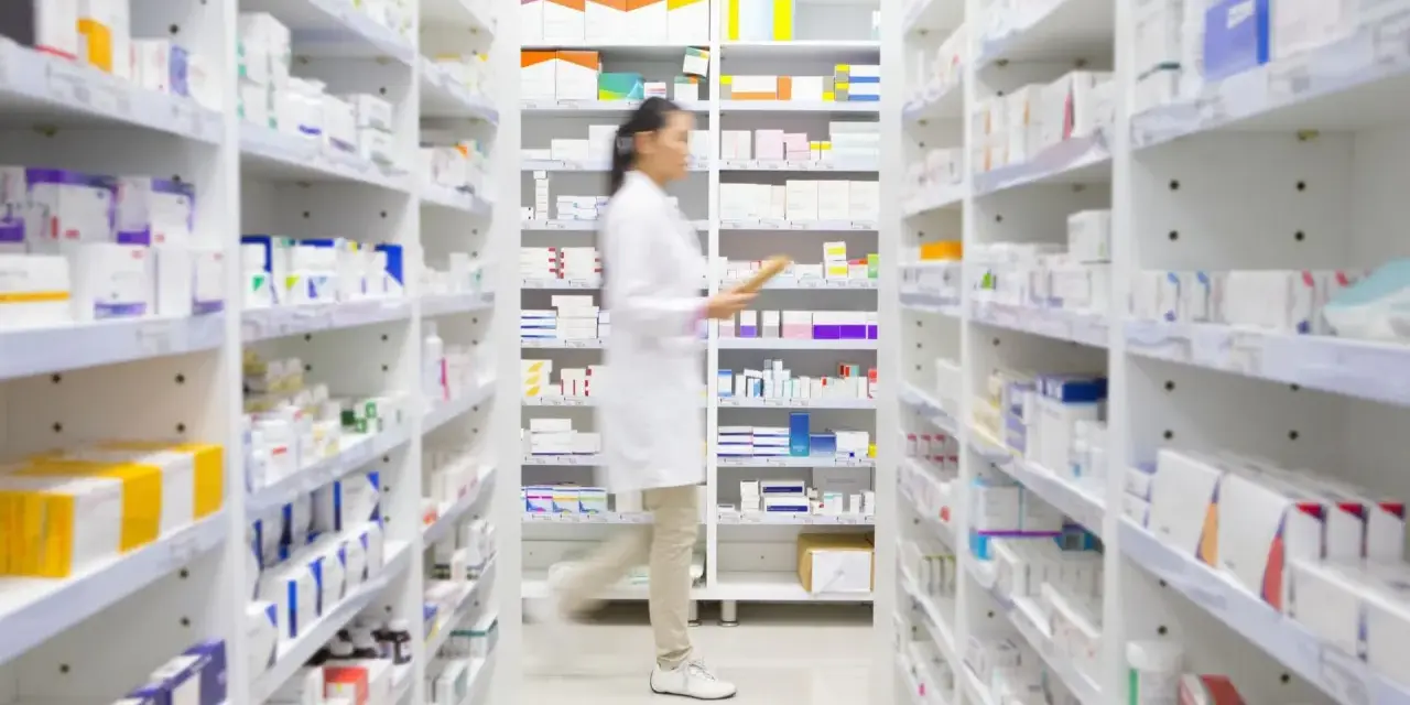 Farmaceutka w białym fartuchu idzie między regałami pełnymi leków, szukając amoksycyliny.