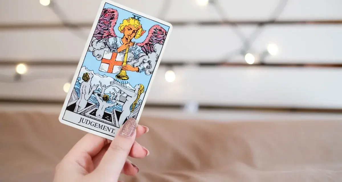 Una mano sostiene la carta de **el juicio tarot**, mostrando un ángel con trompeta y personas resucitando.