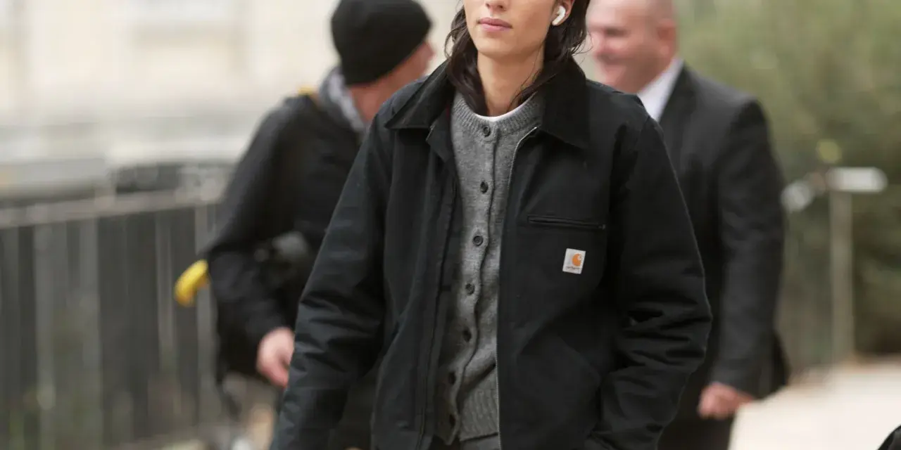 Donna con giacca Carhartt nera e cardigan grigio, cuffiette bianche, cammina con sicurezza.