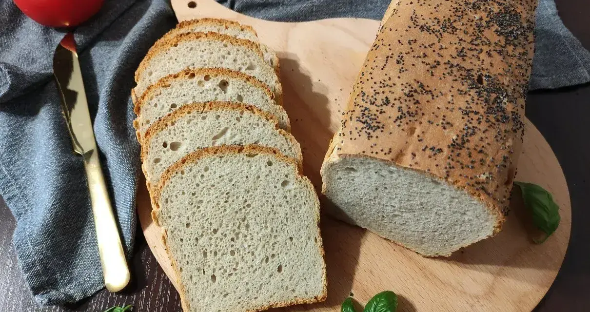 Świeżo upieczony chleb pszenny z makiem, pokrojony w kromki, obok pomidor i złoty nóż.