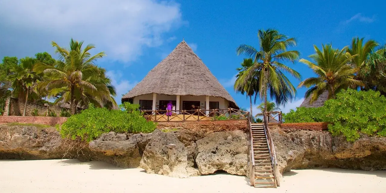 Tropikalny domek z krytym strzechą dachem na skale, obok palm. Marzysz o podróży? Sprawdź, ile się leci na Zanzibar!