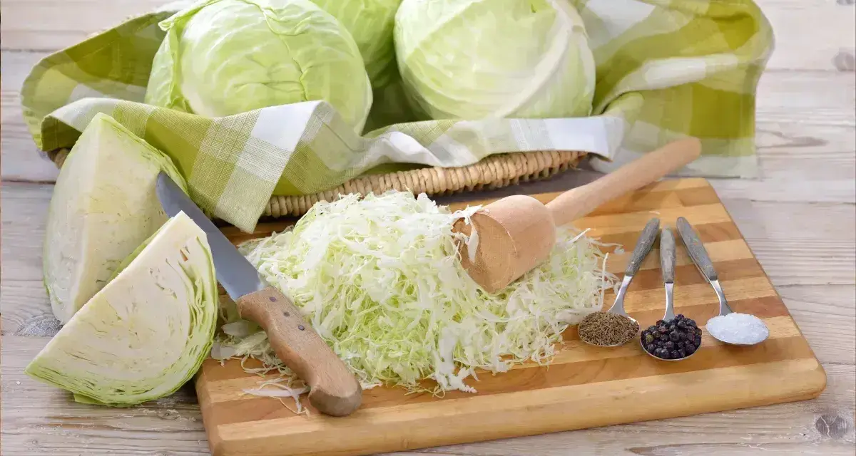 Przygotowanie kiszonej kapusty: ile soli na 10kg? Na desce poszatkowana kapusta, nóż, tłuczek, kminek, pieprz i sól.