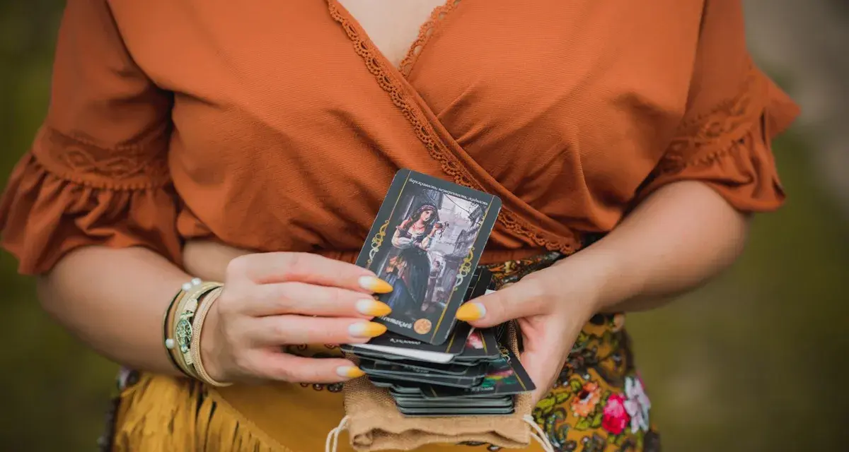 Kobieta w pomarańczowej bluzce trzyma talię kart, na wierzchu widać kartę z cyganką i napisem "3 denary tarot".