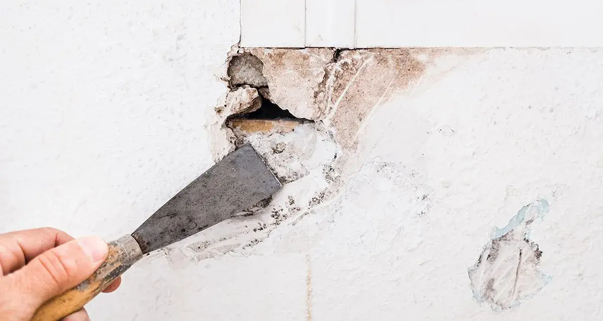 Mano con spatola rimuove intonaco da un buco nel muro troppo grande, rivelando mattoni e detriti.