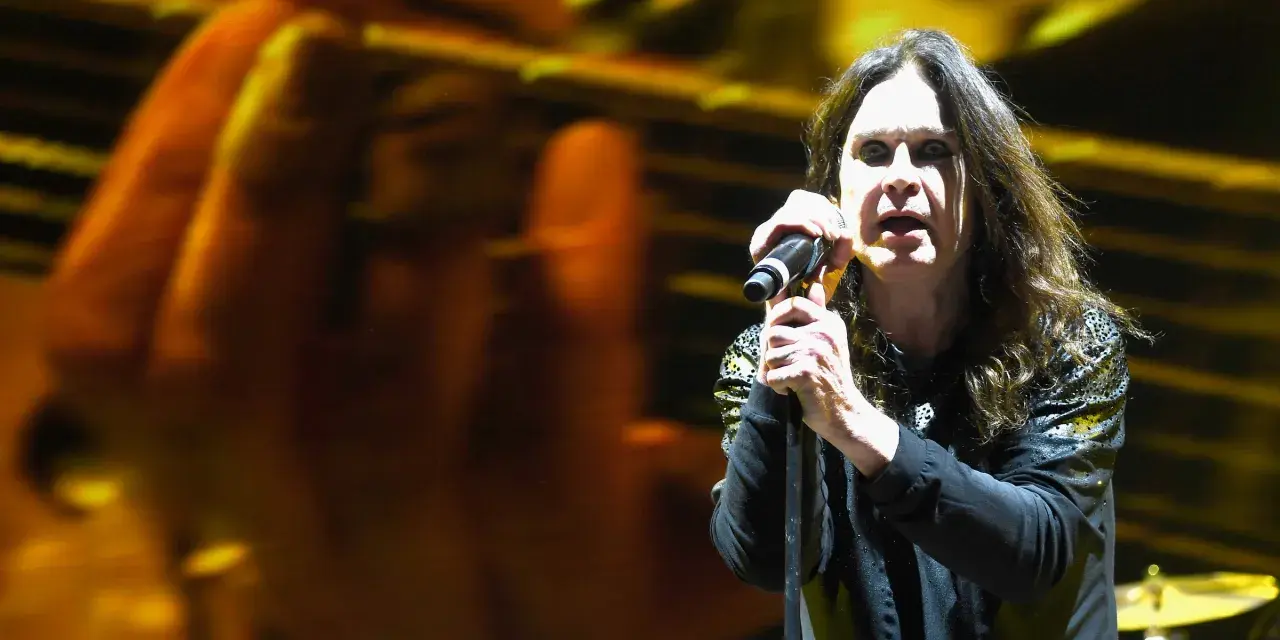 Ozzy Osbourne śpiewa do mikrofonu na koncercie, z rozmytym tłem w kolorach żółci i pomarańczy.