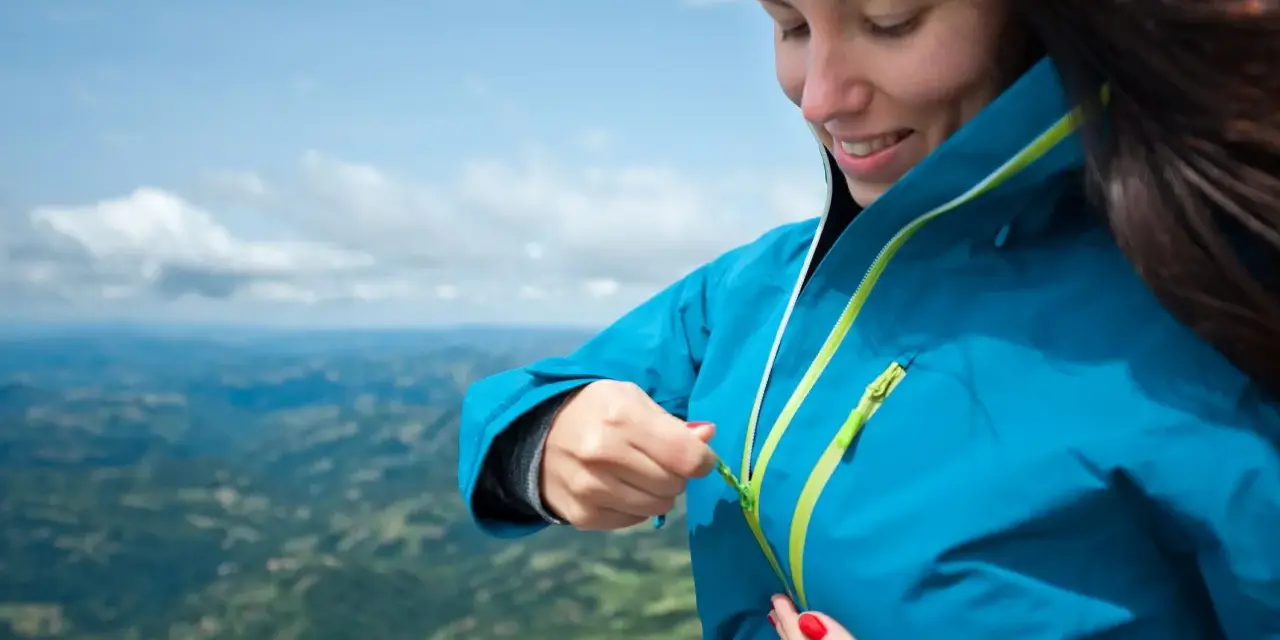 Donna con impermeabile montagna blu, chiude la zip. Paesaggio verdeggiante sotto un cielo azzurro.