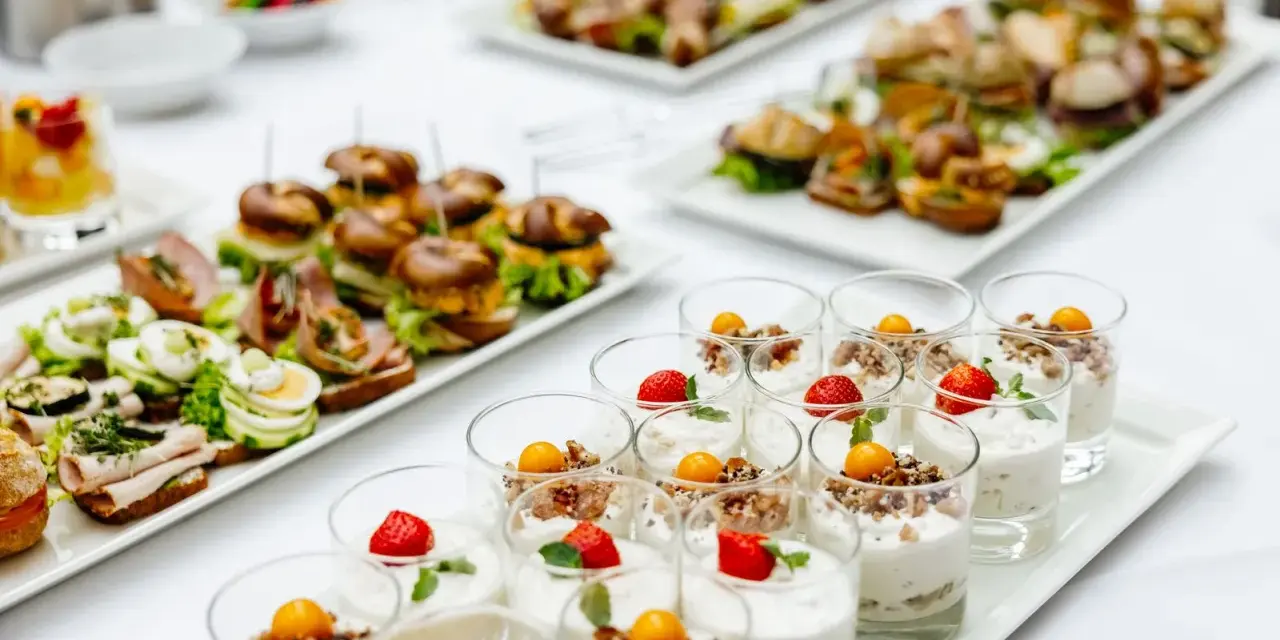 Varietà di antipasti freddi per buffet: mini hamburger, tartine con uova e salumi, e dolcetti in bicchiere con frutta e yogurt.