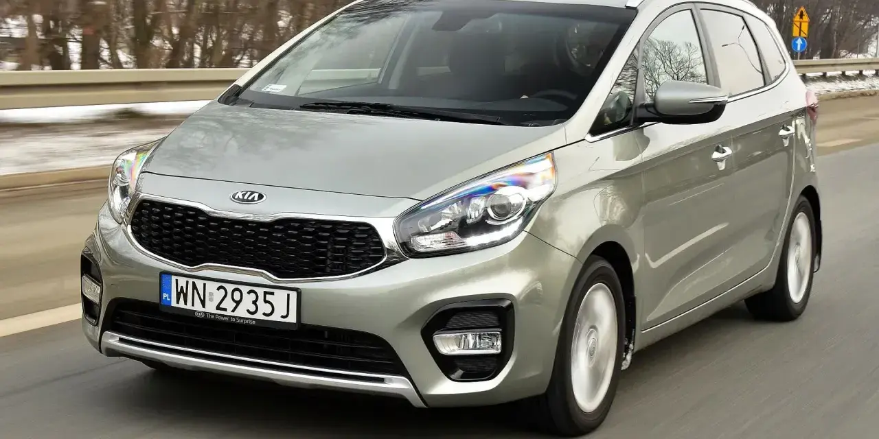 Srebrny Kia Carens, typowy MPV, jedzie po drodze. To auto rodzinne, idealne na wycieczki.