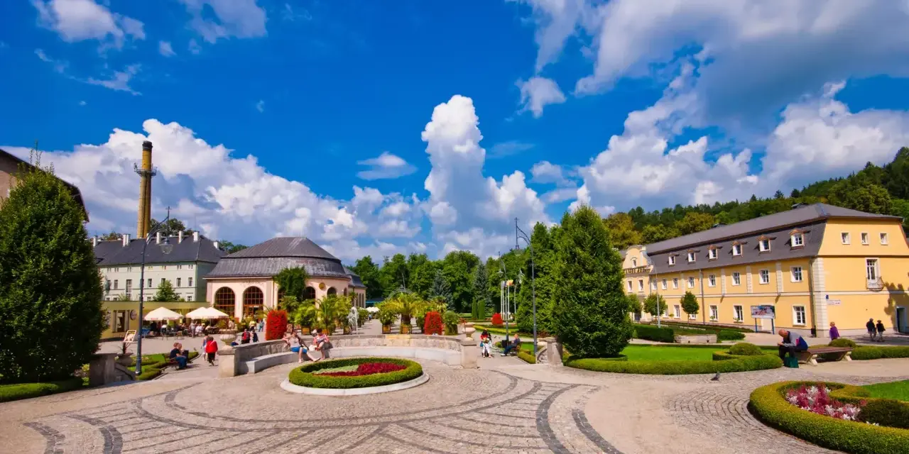 Słoneczny dzień w Kudowie Zdrój. Spacerowicze odpoczywają w pięknym parku, podziwiając architekturę i zieleń. To idealne miejsce na relaks i odkrywanie atrakcji.