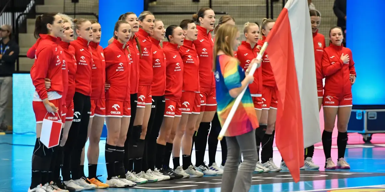 Drużyna piłki ręcznej kobiet Polski w czerwonych strojach stoi w rzędzie, trzymając polską flagę.