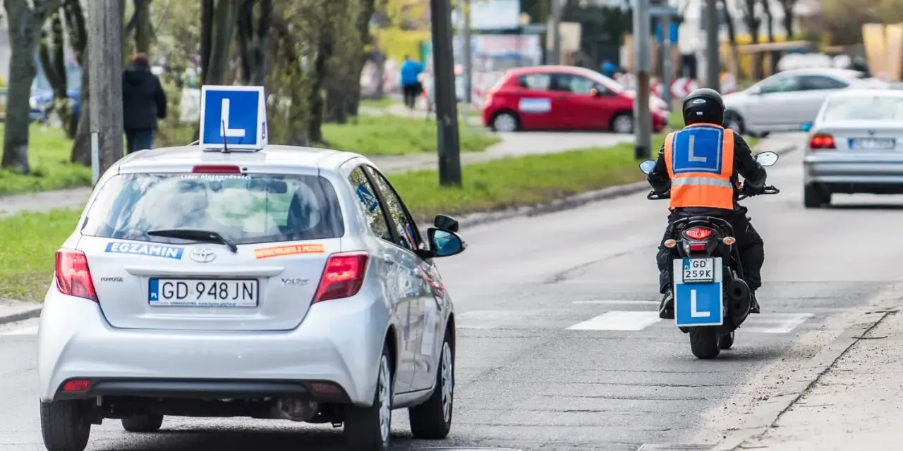 Srebrny samochód z literą "L" na dachu i motocyklista w kamizelce odblaskowej, obaj z literą "L" z tyłu, poruszają się po drodze. To kategoria a nauki jazdy.