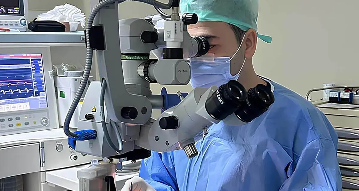 Chirurgo con maschera e camice blu opera con microscopio Zeiss. Monitor vitale e strumentazione indicano un intervento post-operatorio vitrectomia gas.
