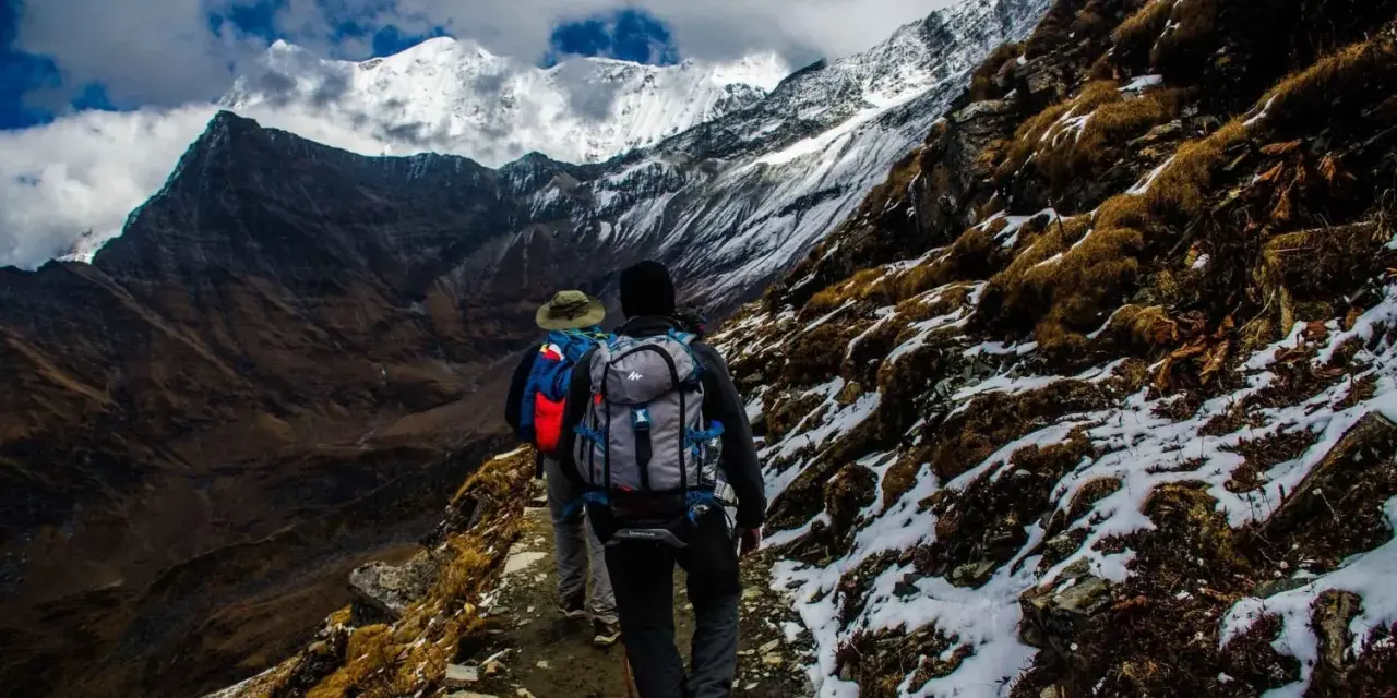 Dwóch turystów z plecakami wędruje ścieżką u podnóża ośnieżonych gór, przypominających krajobraz Mount Kenya.