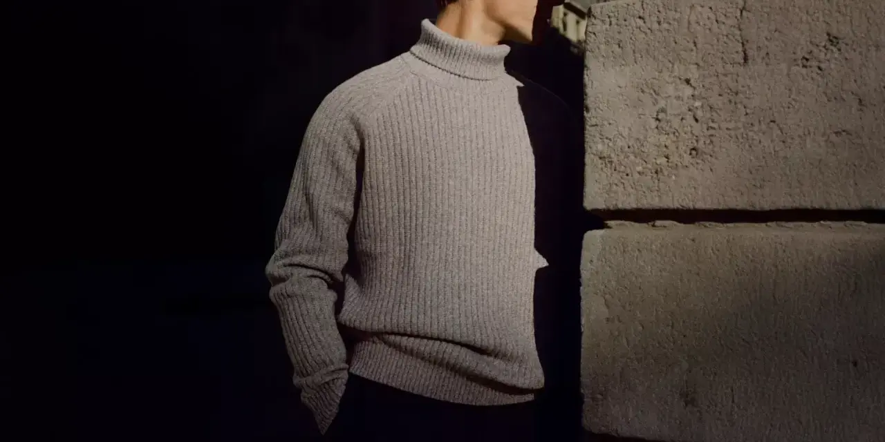 Uomo con un elegante outfit maglione collo alto grigio, in piedi vicino a un muro di pietra.