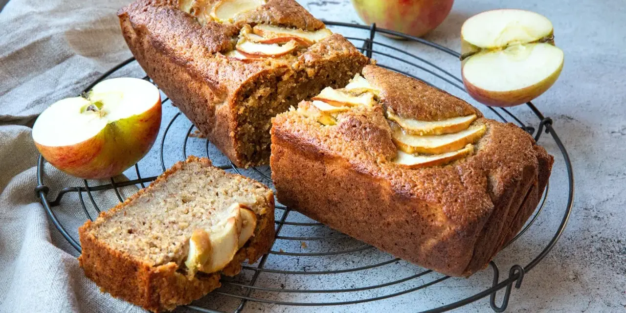 Saftiger Apfelkuchen mit Apfelmus-Note, dekoriert mit frischen Apfelscheiben. Daneben liegen Äpfel und ein Stück Kuchen.