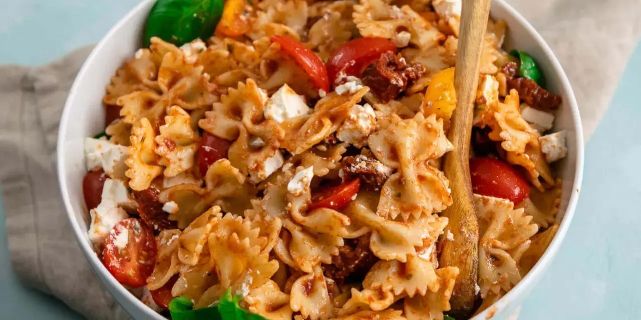 Ein köstlicher, sommerlicher Nudelsalat mit Farfalle, Tomaten, Feta und Basilikum, serviert in einer weißen Schüssel mit Holzutensilien.