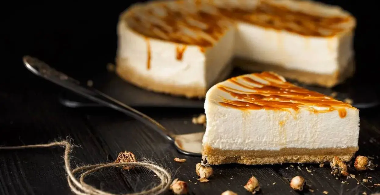 Una fetta di cheesecake senza panna, con glassa caramellata, su un piatto da portata, con la torta intera sullo sfondo.