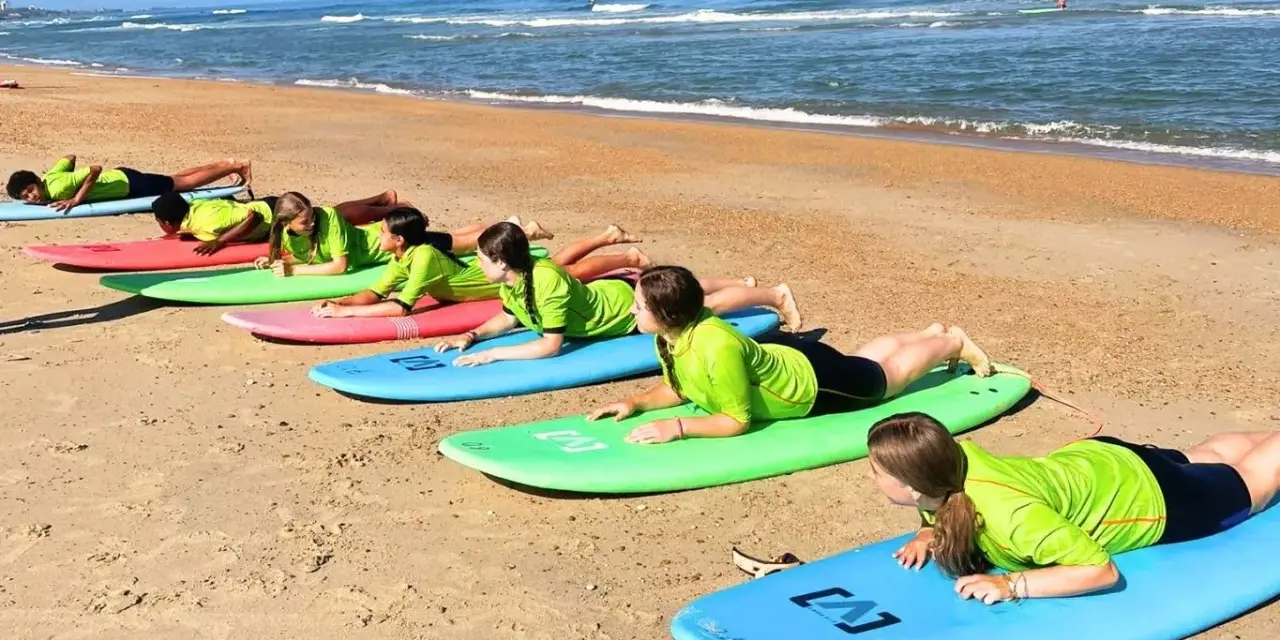 Des adolescents en stage sportif sur la plage, allongés sur des planches de surf, apprenant les bases avant de se jeter à l'eau.