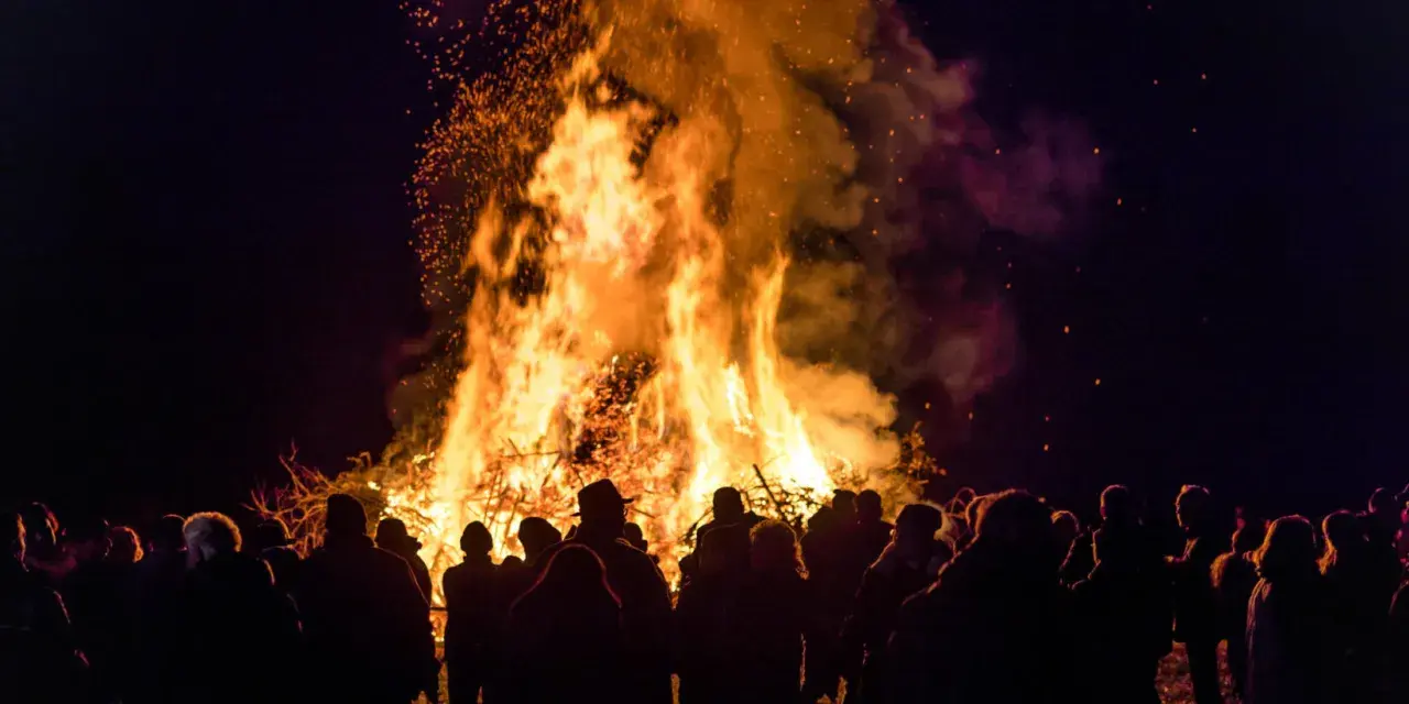 Menschen versammeln sich um ein helles Osterfeuer. Die Flammen lodern hoch in den Nachthimmel, Funken fliegen. Das Osterfeuer symbolisiert die christliche Bedeutung von Licht und Auferstehung.