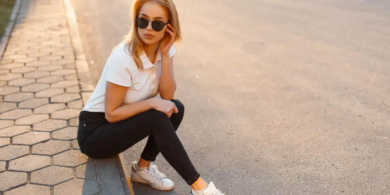 Blondynka w czarnych jeansach i białej koszulce polo, siedząca na chodniku. Stylowa inspiracja, jak nosić koszulka polo damska na co dzień.