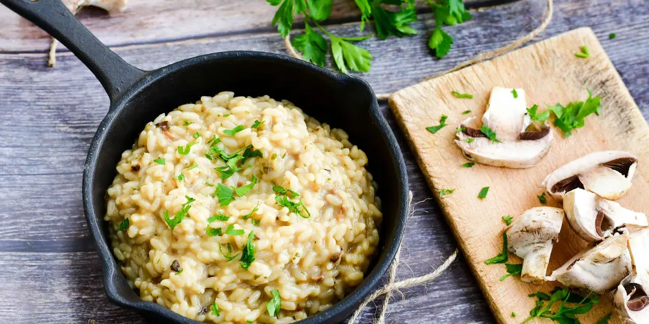 Risotto z grzybami w czarnej patelni, ozdobione natką pietruszki, obok pokrojone pieczarki na desce.