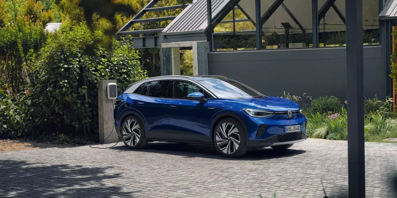 Niebieski Volkswagen ID.4 ładuje się przy domu, otoczony zielenią.