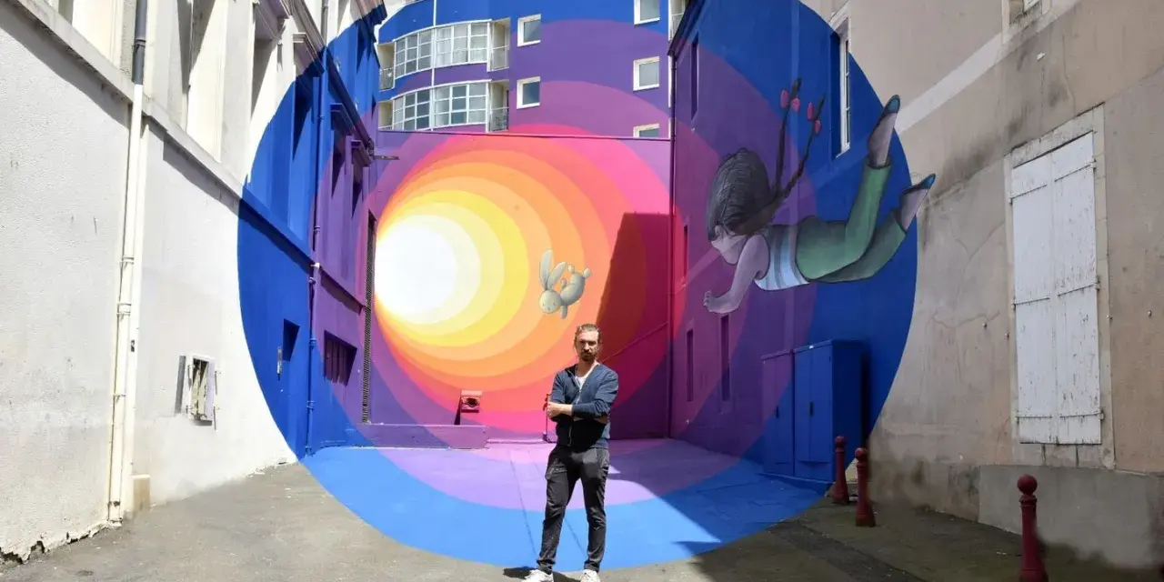 Un artiste street art français pose devant sa fresque colorée d'une fillette et d'un lapin flottant dans un tunnel psychédélique.