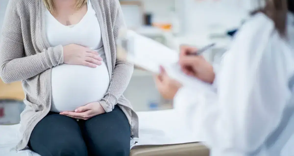 Kobieta w ciąży siedzi na kozetce, trzymając dłonie na brzuchu. Lekarz spisuje dokumenty, prawdopodobnie wystawiając zaświadczenie o ciąży.