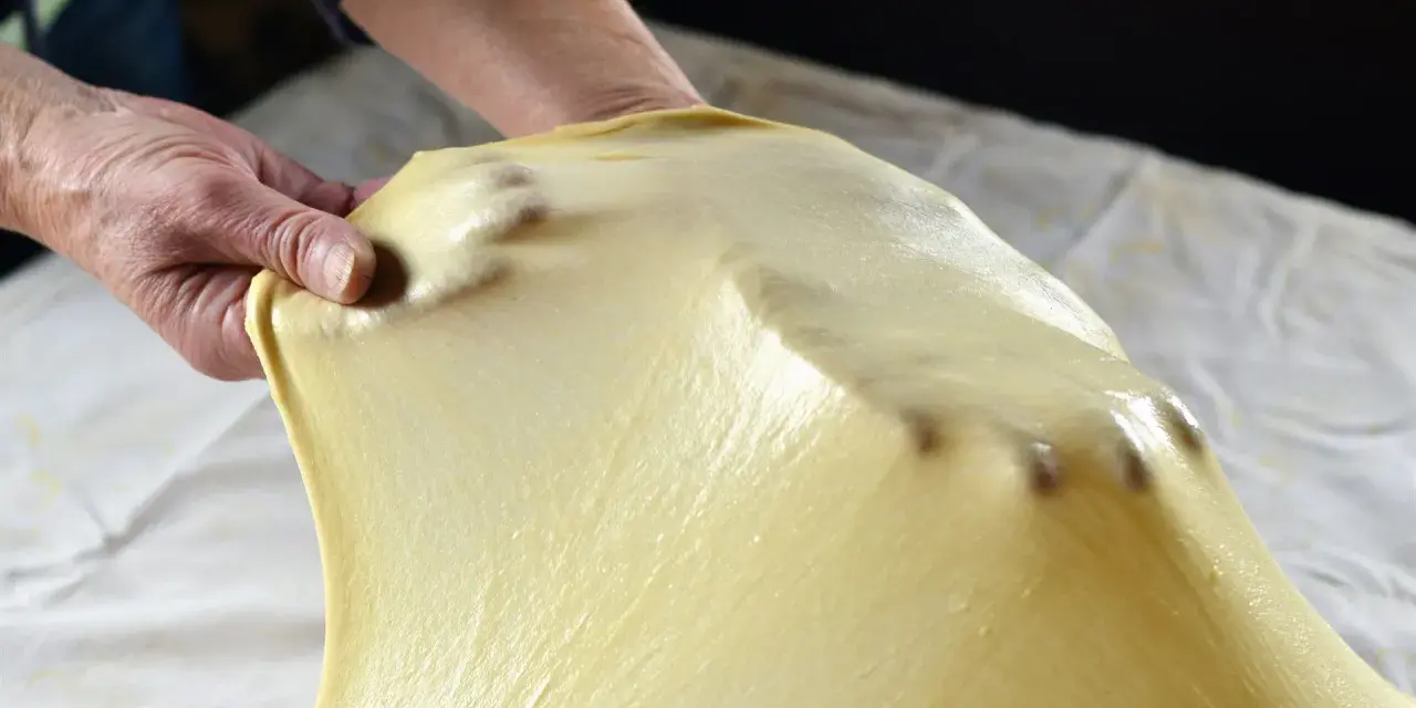 Mani che stendono la pasta matta per una ricetta tradizionale. La pasta è sottile e traslucida, con le dita che creano delle leggere ondulazioni.