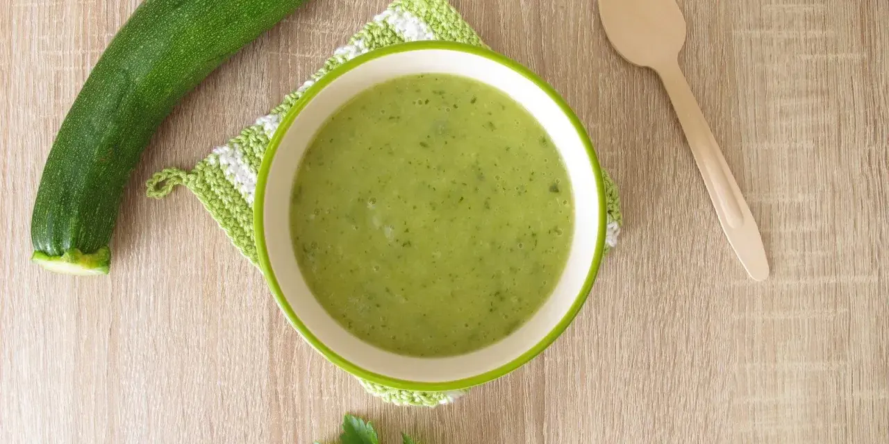Zucchini Brei Baby: Eine Schüssel grüner Zucchinipüree, daneben eine Zucchini und ein Holzlöffel.