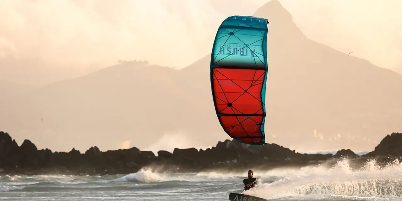 Kitesurfer na desce w akcji, z kolorowym latawcem na tle gór i morza.