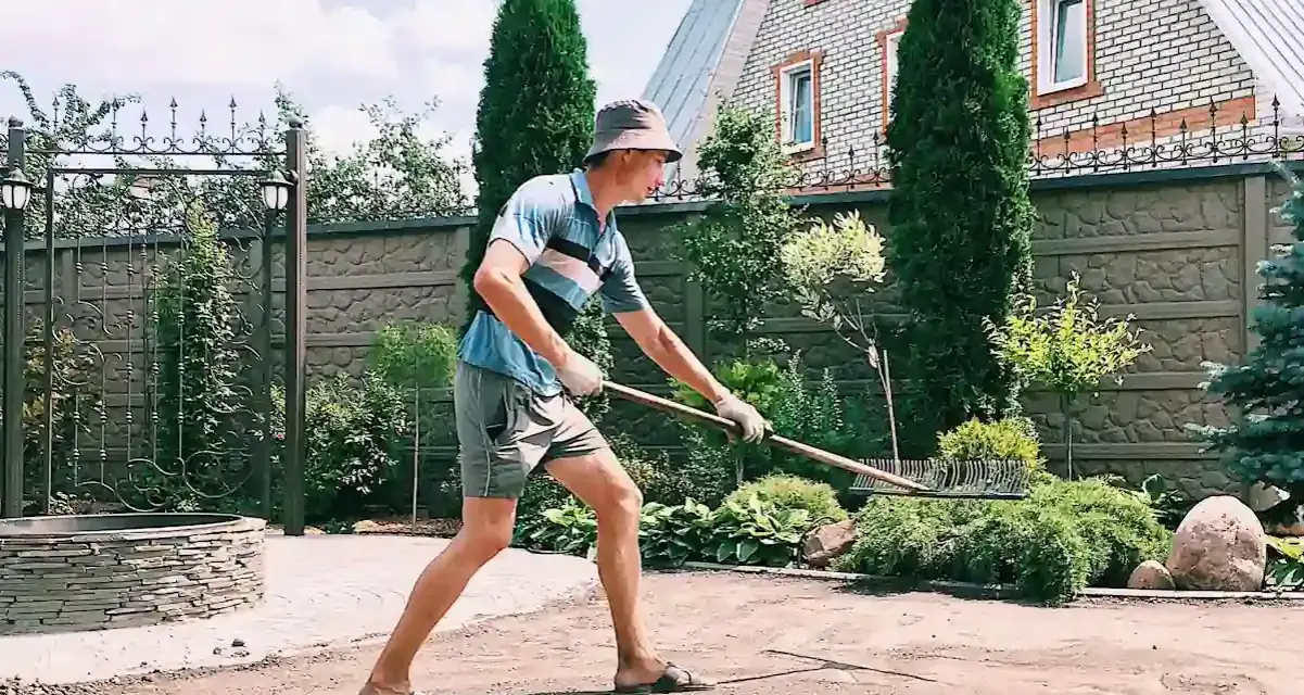 Un homme en chapeau et short utilise un râteau pour faire une place de parking dans son jardin. Le soleil brille sur la pelouse.