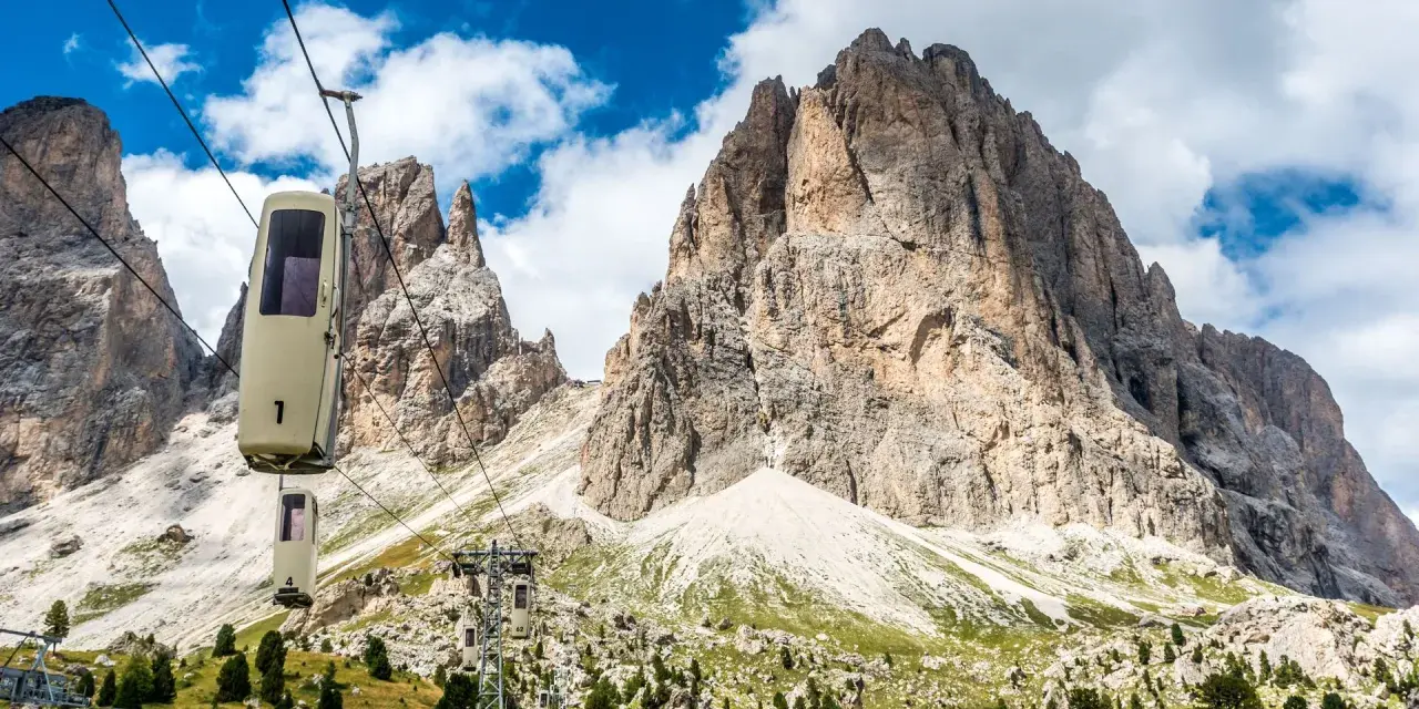 Cabine della funivia salgono verso le imponenti vette del Sassolungo, un anello di roccia maestoso sotto un cielo azzurro con nuvole bianche.