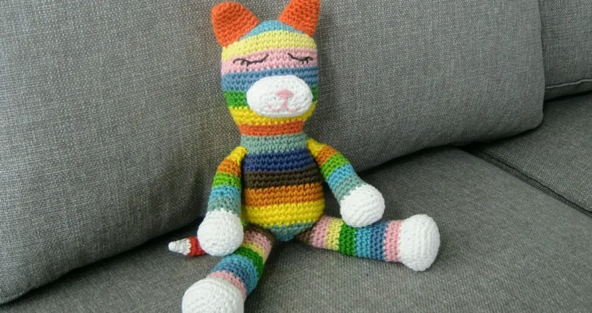 Ett färgglatt virkat gosedjur i form av en katt sitter på en grå soffa.