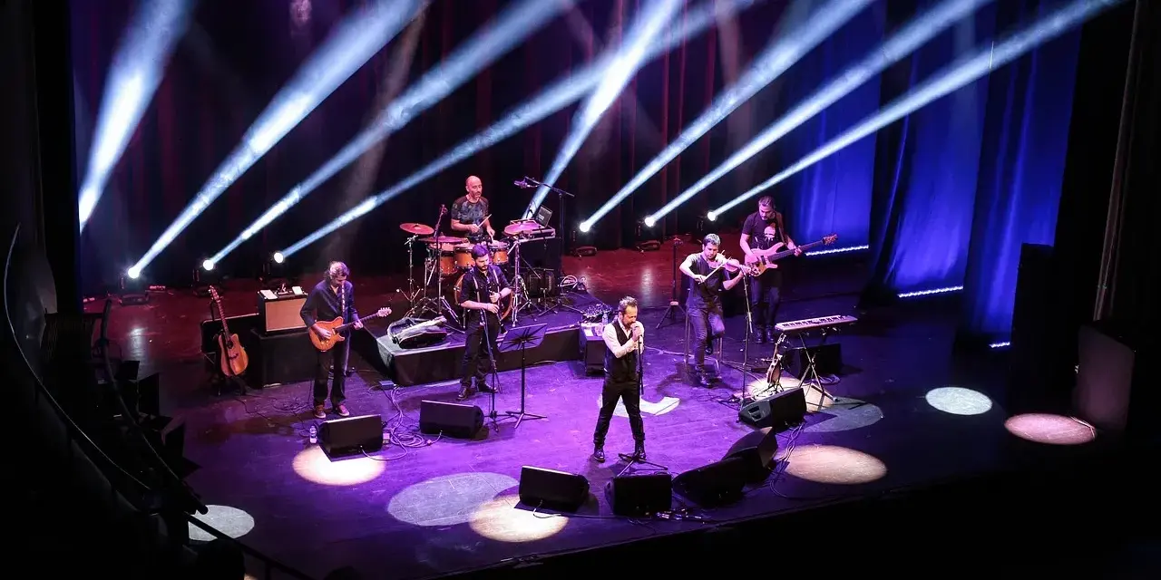 Zespół muzyczny na scenie, oświetlony niebieskimi i fioletowymi światłami. Wokalista na pierwszym planie, gitarzyści, basista, skrzypek i perkusista. Jak liczyć takty w muzyce, można się nauczyć, obserwując ich rytm.