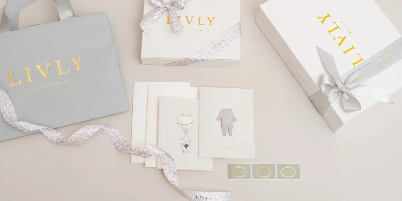En fin babyshower present tjej med en grå presentpåse, vita presentaskar med guldig text och leopardmönstrat band, samt kort med illustrationer av en elefant i ballong och en babybody.