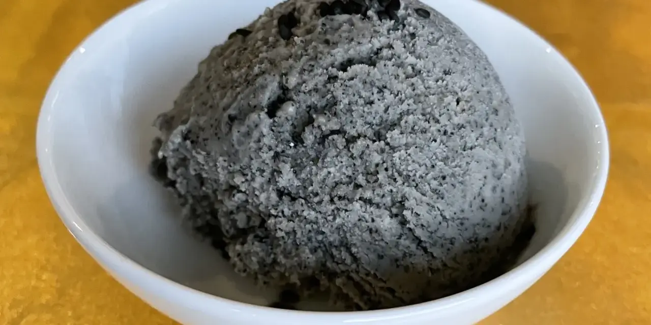 Ein Löffel cremiges Sesam Eis, garniert mit schwarzen Sesamkörnern, in einer weißen Schale.