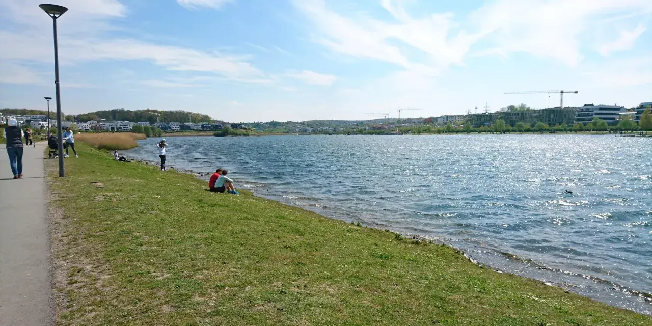 Ein sonniger Tag am Dortmunder See. Menschen spazieren am Ufer entlang, während ein Paar auf dem Gras sitzt und die Aussicht genießt.