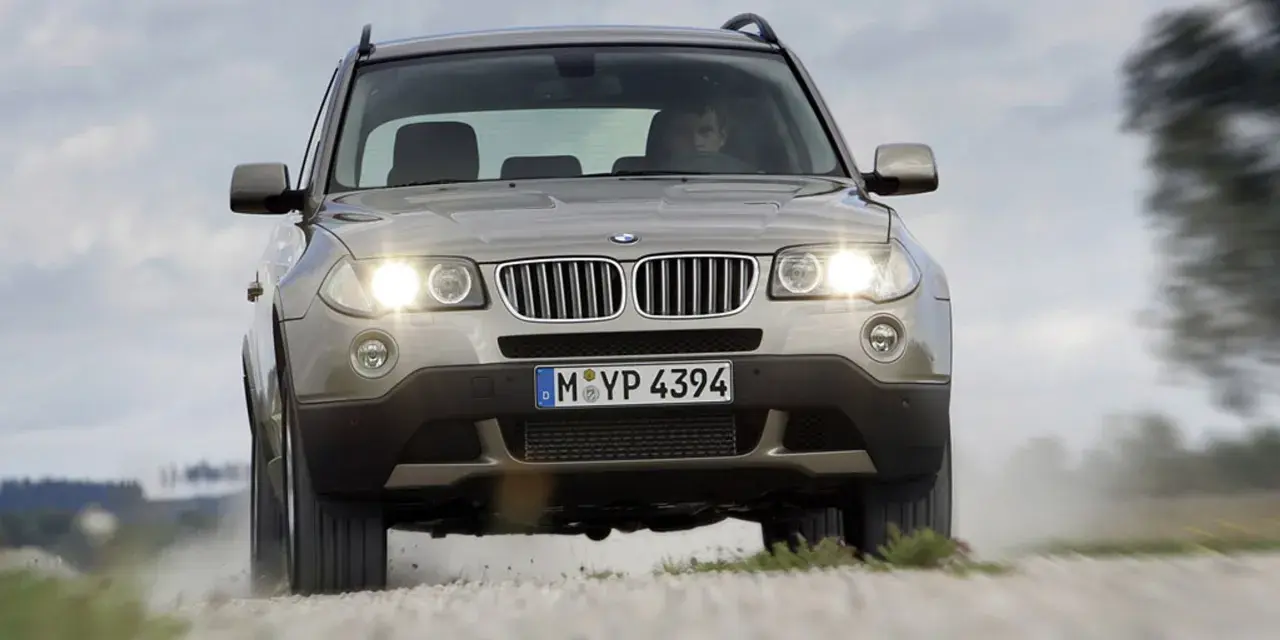 Złoty SUV BMW X3 z włączonymi światłami jedzie po szutrowej drodze, unosząc kurz. To jeden z najbardziej komfortowych aut na drodze.