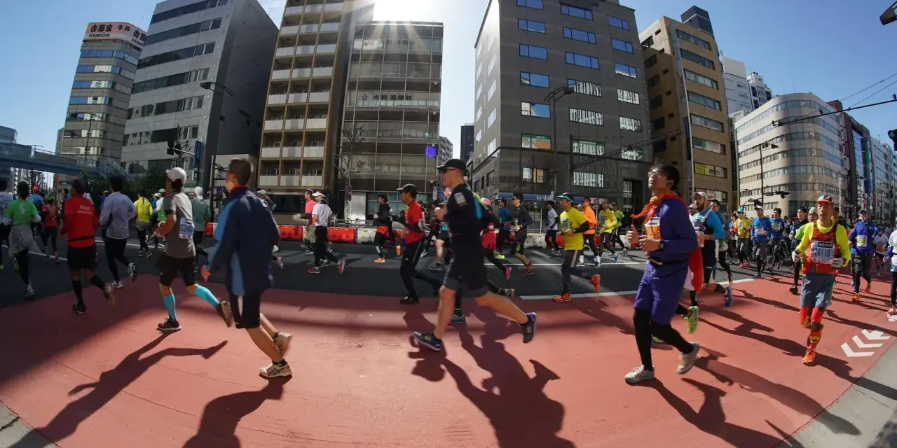 Atleti corrono nella maratona di Tokyo, circondati da grattacieli moderni sotto un sole splendente.