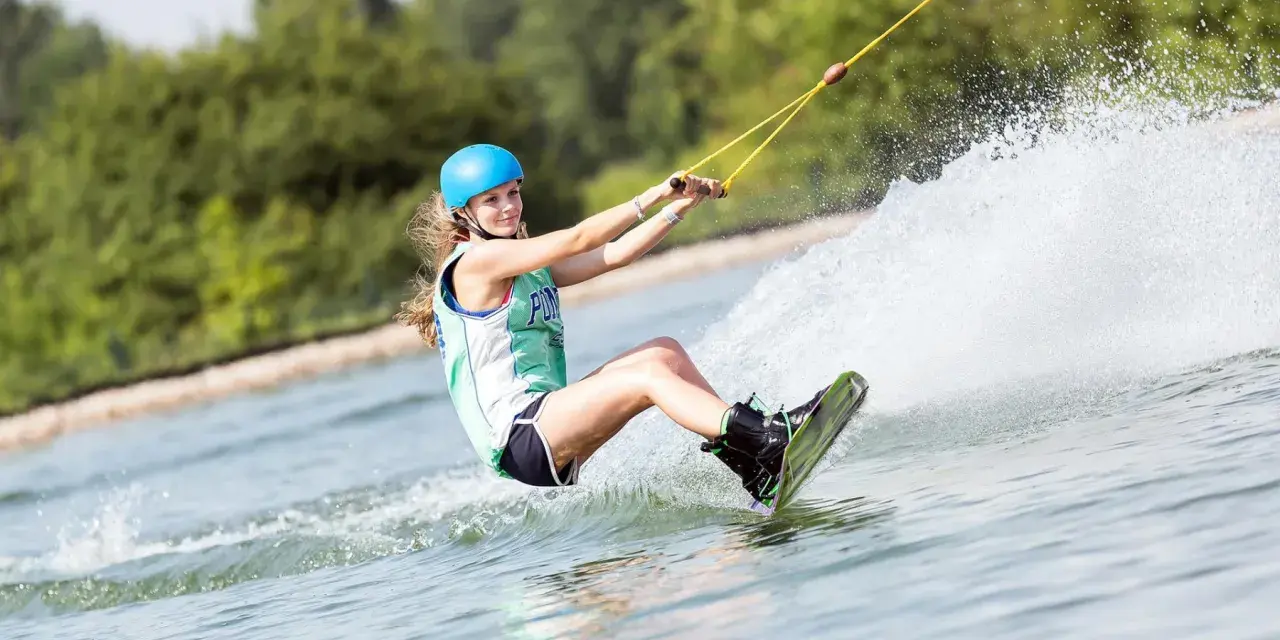 Dziewczyna w kasku i kamizelce pływa na wakeboardzie, tworząc wielki szelment wody.