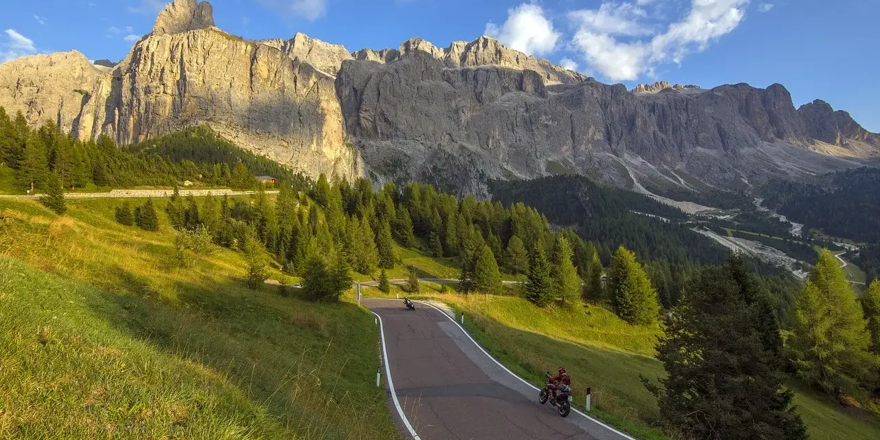 Un motociclista percorre una strada panoramica tra prati verdi e boschi di conifere, con le maestose Dolomiti sullo sfondo. Un'avventura indimenticabile in moto.