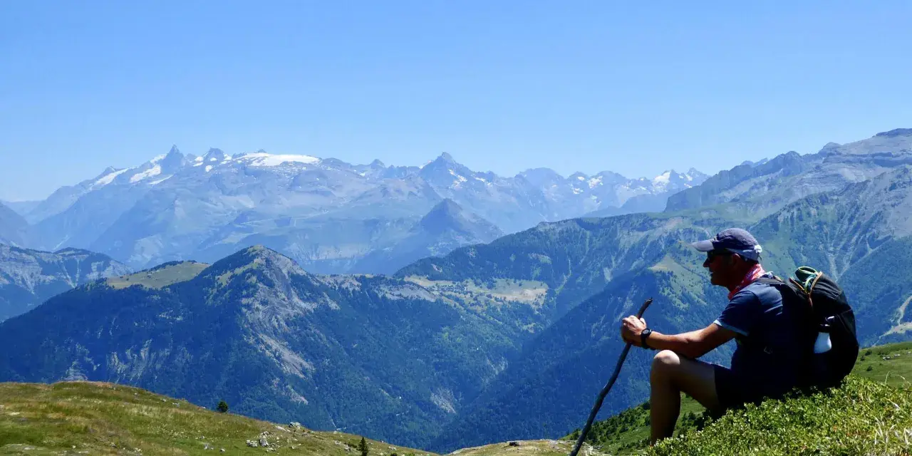 Un randonneur léger profite d'une vue panoramique sur les montagnes. Il est assis sur une colline verdoyante, bâton à la main, admirant le paysage majestueux.