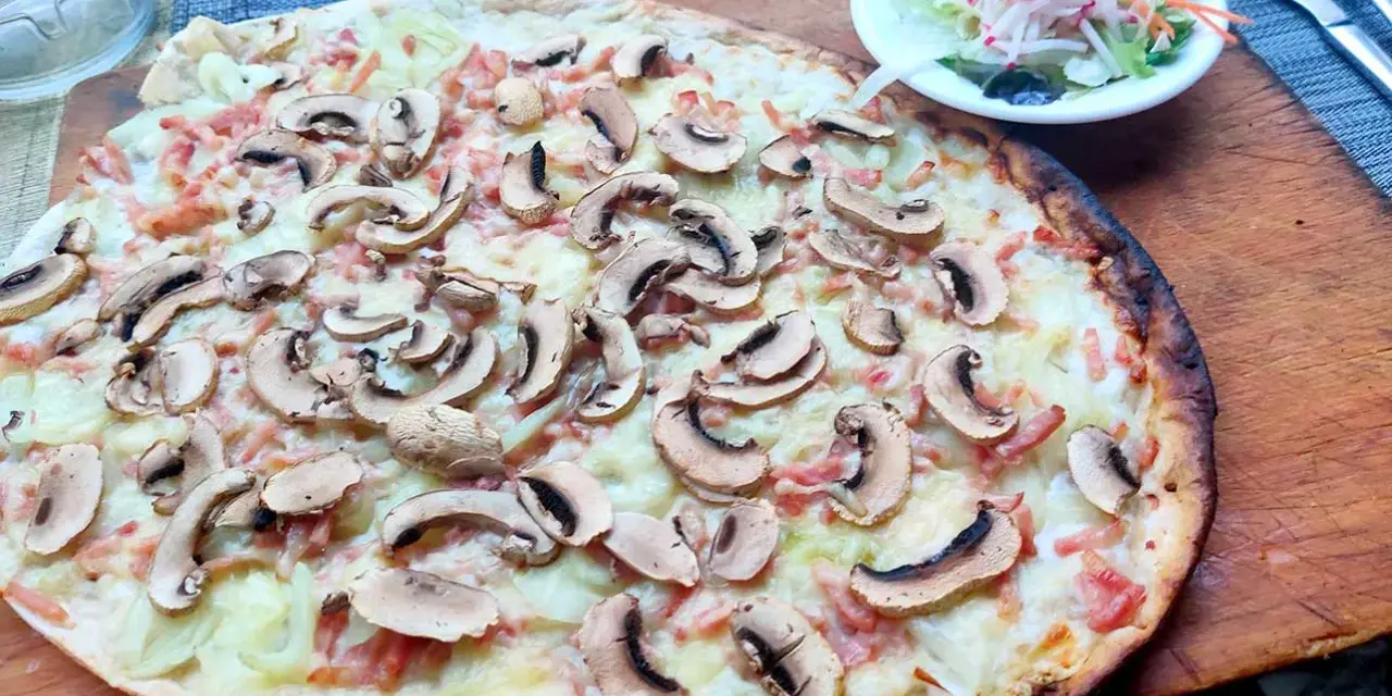 Tarta flambeada con champiñones y bacon, servida sobre tabla de madera. Una pequeña ensalada acompaña.