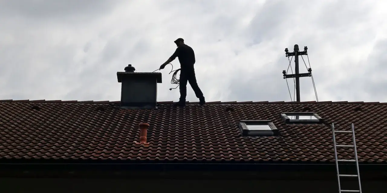 Kominiarz na dachu sprawdza komin. Zastanawiasz się, ile kosztuje przegląd kominiarski?