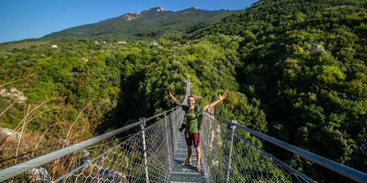 Uomo felice sul laviano ponte tibetano, con montagne verdi e cielo azzurro sullo sfondo.