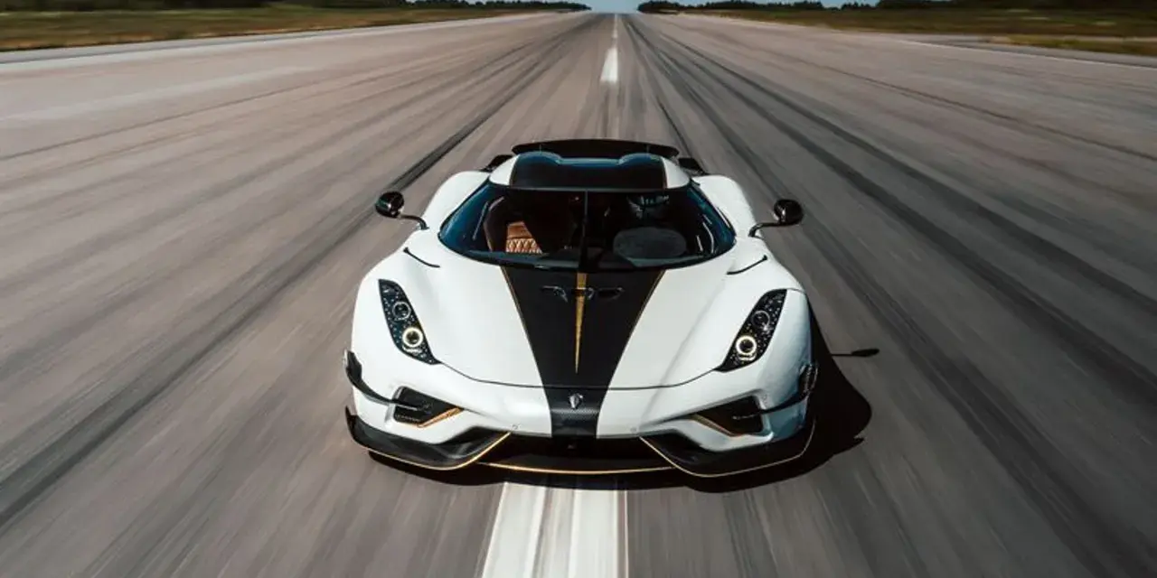 Biały Koenigsegg Regera z czarnym pasem i złotymi akcentami pędzi po pasie startowym, zostawiając za sobą smugę prędkości.