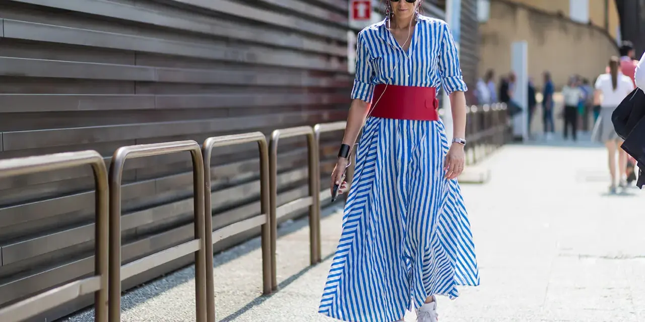 Donna con vestito a righe blu e bianche, cintura rossa e sneakers bianche cammina per strada.